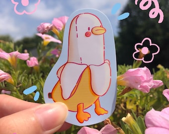 May include: A sticker depicting a cartoon duck-banana hybrid. The design features a yellow banana peel body, a white banana fruit head, and orange feet and beak. The sticker is held against a backdrop of pink flowers and a blue sky.