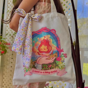 May include: White canvas tote bag featuring a colorful illustration of a flame with a smiling face, held in a hand. The text "A heart is a heavy burden" is below the image. A floral patterned scarf is tied to the handle.
