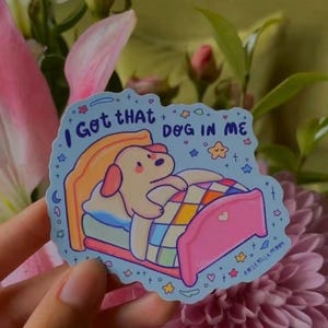 May include: A light blue sticker featuring a cartoon dog tucked into a pink bed with a colorful checkered blanket. The text "I Got THAT DOG IN ME" is printed in dark blue above the dog, surrounded by small stars, hearts, and moon icons.