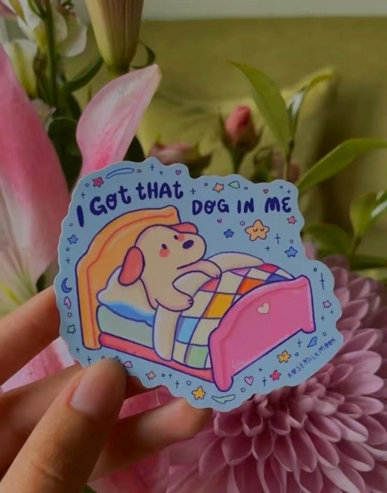May include: A light blue sticker featuring a cartoon dog tucked into a pink bed with a colorful checkered blanket. The text "I Got THAT DOG IN ME" is printed in dark blue above the dog, surrounded by small stars, hearts, and moon icons.