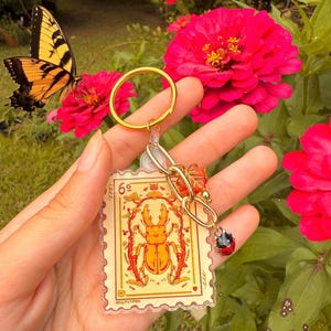 May include: A gold-colored keychain featuring a clear, rectangular charm with a yellow and orange beetle illustration resembling a postage stamp. The keychain includes a gold ring, a chain, and a ladybug charm. A yellow and black butterfly and pink flowers are in the background.