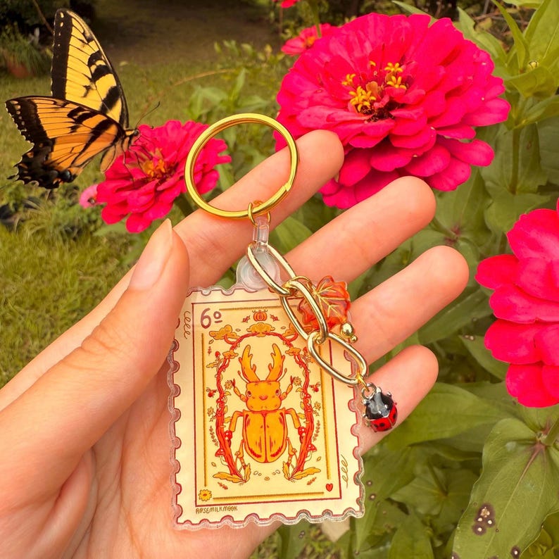 May include: A gold-colored keychain featuring a clear, rectangular charm with a yellow and orange beetle illustration resembling a postage stamp. The keychain includes a gold ring, a chain, and a ladybug charm. A yellow and black butterfly and pink flowers are in the background.