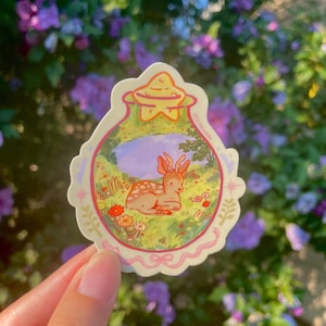 May include: A sticker featuring a cartoon deer resting in a meadow. The sticker has a light pink border with a star-shaped top. The deer is brown with white spots, and the meadow has flowers and green grass.