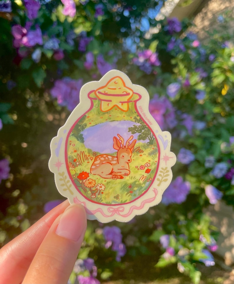 May include: A sticker featuring a cartoon deer resting in a meadow. The sticker has a light pink border with a star-shaped top. The deer is brown with white spots, and the meadow has flowers and green grass.