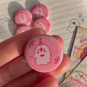 May include: Pink button with a cartoon ghost wearing a party hat and bow tie. The ghost is surrounded by confetti and streamers. The button is 1 inch in diameter.
