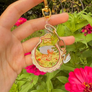 May include: A clear acrylic keychain with a gold clasp and chain. The keychain features a whimsical illustration of a deer in a meadow, a swan charm, and a small leaf charm. The background includes pink flowers and green foliage.