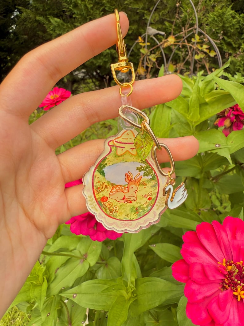 May include: A clear acrylic keychain with a gold clasp and chain. The keychain features a whimsical illustration of a deer in a meadow, a swan charm, and a small leaf charm. The background includes pink flowers and green foliage.