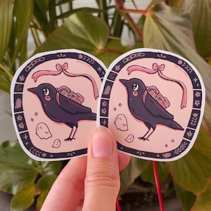 May include: Two black crow stickers with pink ribbons and small ghost illustrations. The crows are carrying small bags with pink accents. The stickers have a black and white border with a floral design.