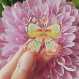 May include: A clear, decorative butterfly-shaped pin with yellow, orange, and green accents. The pin features a detailed design with a flower and star details. The pin is held in front of a pink flower.