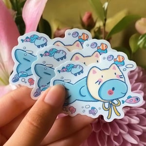 May include: A stack of stickers featuring a cartoon cat in a blue and yellow diving suit, surrounded by fish and bubbles. The stickers have a white border and are held in a hand against a background of flowers.