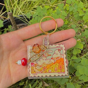May include: A clear acrylic keychain with a gold-colored ring and chain. The keychain features a colorful illustration resembling a postage stamp, with a landscape scene and the number "15". A red and white bead charm hangs from the chain.