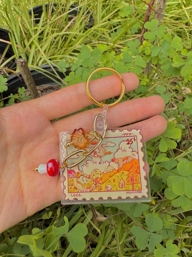 May include: A clear acrylic keychain with a gold-colored ring and chain. The keychain features a colorful illustration resembling a postage stamp, with a landscape scene and the number "15". A red and white bead charm hangs from the chain.