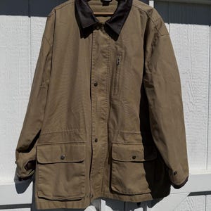 May include: A tan utility jacket featuring a dark brown leather collar, a vertical zippered chest pocket, and two large front patch pockets with snap closures. The jacket has a button-down front placket and long sleeves with dark brown leather cuffs.