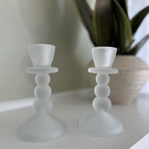 May include: Two frosted glass candle holders with a beaded design. The holders are white and have a round base.
