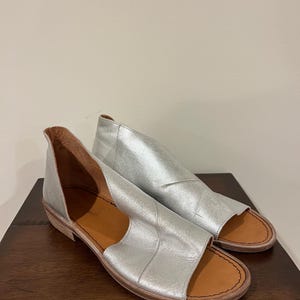 Puede incluir: Un par de sandalias de cuero plateado con punta abierta y tac&oacute;n bajo. Las sandalias tienen un dise&ntilde;o recortado en los laterales y una plantilla marr&oacute;n. Los zapatos se exhiben sobre una superficie de madera marr&oacute;n oscuro.