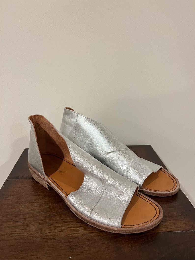 Puede incluir: Un par de sandalias de cuero plateado con punta abierta y tac&oacute;n bajo. Las sandalias tienen un dise&ntilde;o recortado en los laterales y una plantilla marr&oacute;n. Los zapatos se exhiben sobre una superficie de madera marr&oacute;n oscuro.