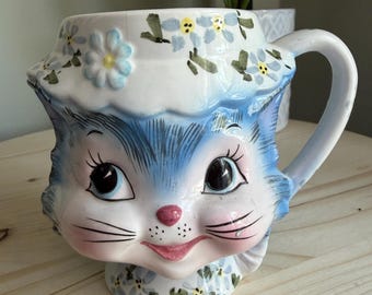 Könnte beinhalten: Ein Keramikbecher in Form eines Katzenkopfes mit blauem Fell, großen schwarzen Augen, einer rosa Nase und Schnurrhaaren. Die Katze trägt eine weiße Haube, die mit kleinen blauen und gelben Blumen verziert ist. Der Becher hat einen Henkel und Fuß.