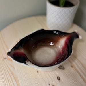 May include: A ceramic serving bowl with a unique leaf-like shape. The bowl features a gradient of colors, transitioning from a dark brown rim to a lighter beige interior, with red accents. The bowl is placed on a light wood surface.