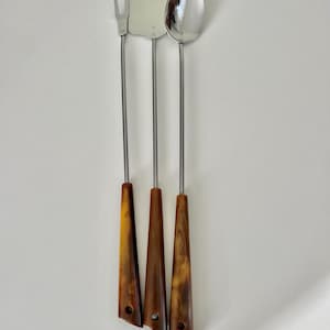 May include: A set of three stainless steel grilling tools: a fork, a spatula, and a spoon. Each tool has a long, thin metal handle and a brown, triangular handle with a hole at the base. The tools are arranged vertically.