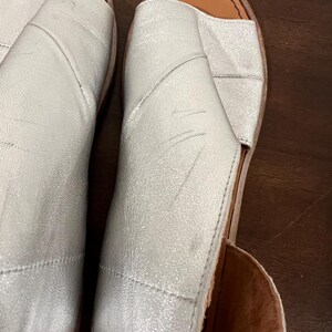 Puede incluir: Par de sandalias plateadas con punta abierta. Las sandalias tienen un brillo met&aacute;lico y un dise&ntilde;o cosido. Las suelas son de color marr&oacute;n claro. Los zapatos parecen ser de cuero.