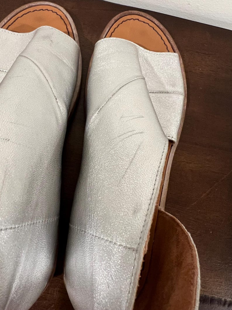 Puede incluir: Par de sandalias plateadas con punta abierta. Las sandalias tienen un brillo met&aacute;lico y un dise&ntilde;o cosido. Las suelas son de color marr&oacute;n claro. Los zapatos parecen ser de cuero.