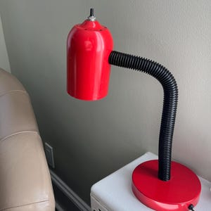 May include: A red desk lamp with a flexible black neck and a round red base. The lamp has a cylindrical shade and a small on/off switch on top. The lamp is sitting on a white surface.