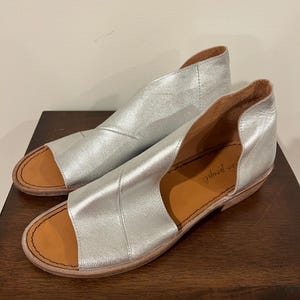 Puede incluir: Un par de sandalias de cuero plateado con punta abierta y tac&oacute;n bajo. Las sandalias tienen un dise&ntilde;o recortado en la parte superior y una plantilla marr&oacute;n. La marca "free people" es visible en la plantilla.