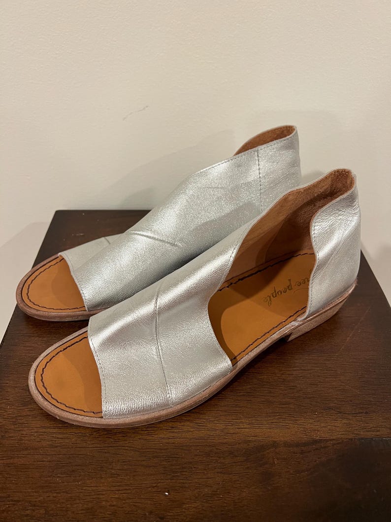 Puede incluir: Un par de sandalias de cuero plateado con punta abierta y tac&oacute;n bajo. Las sandalias tienen un dise&ntilde;o recortado en la parte superior y una plantilla marr&oacute;n. La marca "free people" es visible en la plantilla.