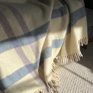 May include: A soft, cream-colored blanket with blue and lavender stripes. The blanket has a fringed edge and is draped over a surface, with sunlight illuminating the fabric.