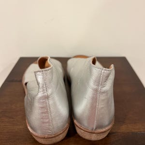 Puede incluir: Un par de bailarinas de cuero plateado con punta redondeada y suela baja marr&oacute;n. Los zapatos se exhiben sobre una superficie de madera marr&oacute;n oscuro sobre un fondo blanco liso. Dise&ntilde;o sencillo y minimalista.