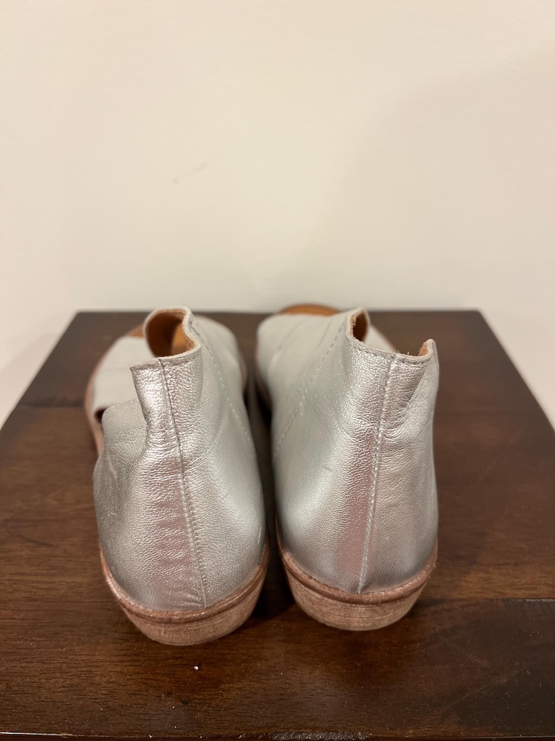 Puede incluir: Un par de bailarinas de cuero plateado con punta redondeada y suela baja marr&oacute;n. Los zapatos se exhiben sobre una superficie de madera marr&oacute;n oscuro sobre un fondo blanco liso. Dise&ntilde;o sencillo y minimalista.