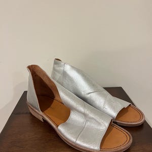 Puede incluir: Un par de sandalias de cuero plateado con punta abierta y tac&oacute;n bajo. Las sandalias tienen un dise&ntilde;o recortado en los laterales y una suela marr&oacute;n. Los zapatos se exhiben sobre una superficie de madera oscura.