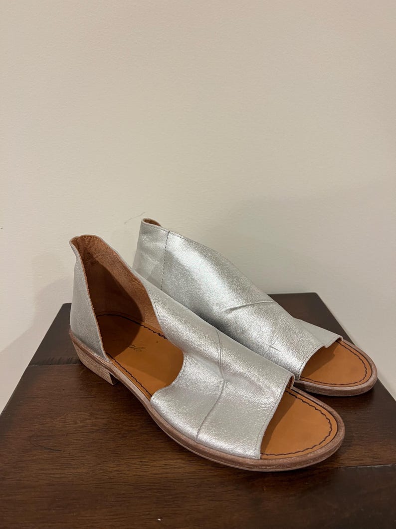 Puede incluir: Un par de sandalias de cuero plateado con punta abierta y tac&oacute;n bajo. Las sandalias tienen un dise&ntilde;o recortado en los laterales y una suela marr&oacute;n. Los zapatos se exhiben sobre una superficie de madera oscura.