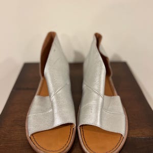 Puede incluir: Un par de sandalias de cuero plateado con punta abierta y dise&ntilde;o recortado. Las sandalias tienen una plantilla de cuero marr&oacute;n y un borde cosido. Los zapatos se exhiben sobre una superficie de madera marr&oacute;n oscuro.
