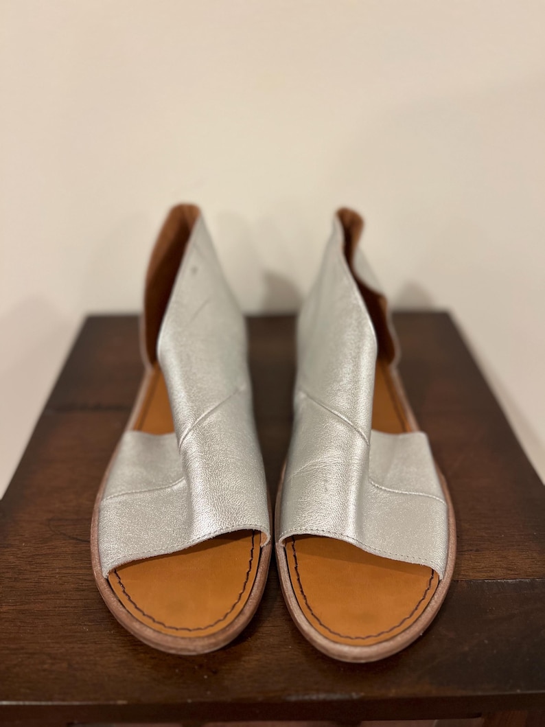 Puede incluir: Un par de sandalias de cuero plateado con punta abierta y dise&ntilde;o recortado. Las sandalias tienen una plantilla de cuero marr&oacute;n y un borde cosido. Los zapatos se exhiben sobre una superficie de madera marr&oacute;n oscuro.