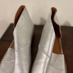 Puede incluir: Un par de sandalias de cuero plateado con punta abierta y dise&ntilde;o recortado. Las sandalias tienen una plantilla de cuero marr&oacute;n y una suela plana. Las sandalias se exhiben sobre una superficie marr&oacute;n oscuro.