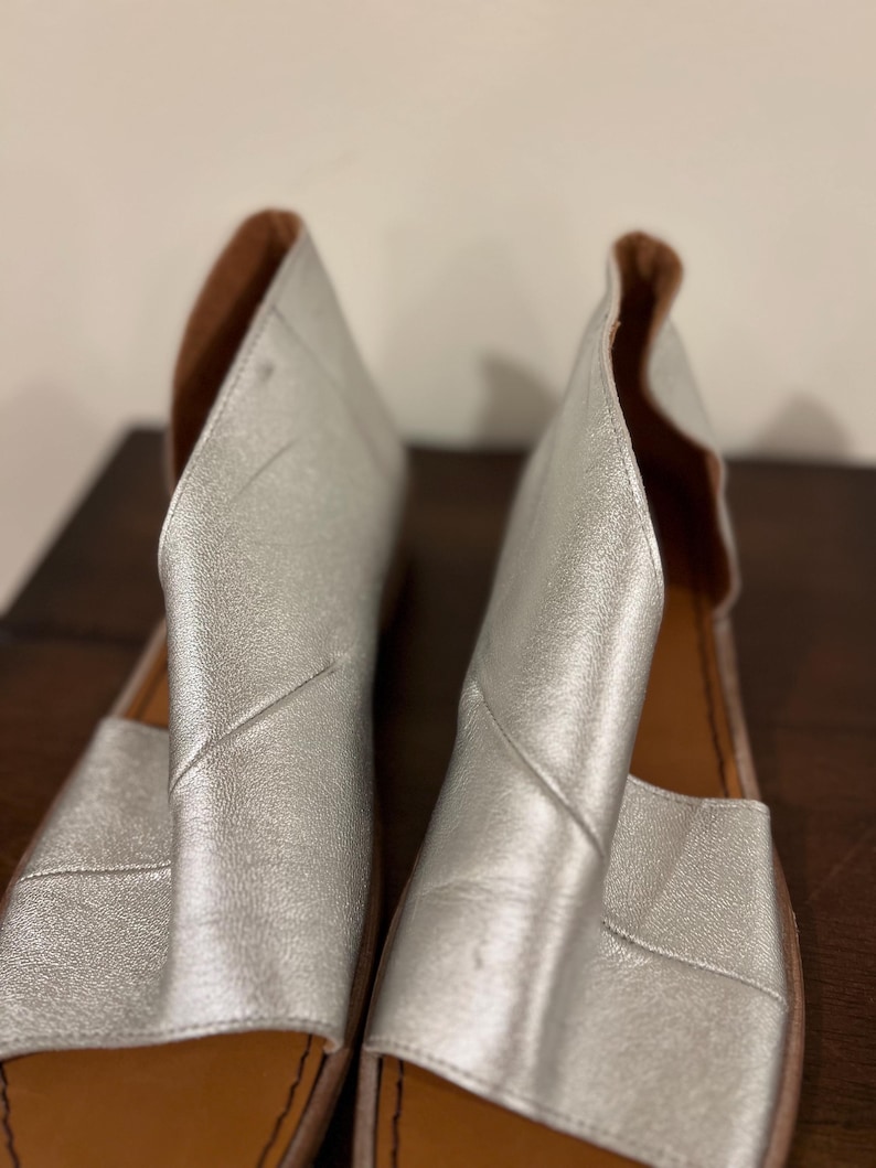 Puede incluir: Un par de sandalias de cuero plateado con punta abierta y dise&ntilde;o recortado. Las sandalias tienen una plantilla de cuero marr&oacute;n y una suela plana. Las sandalias se exhiben sobre una superficie marr&oacute;n oscuro.