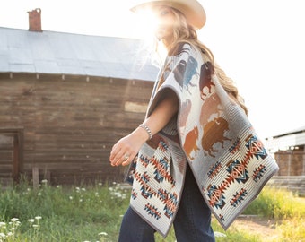 Puede incluir: Una mujer con un sombrero de vaquero blanco y un poncho estampado en gris y marrón camina por un campo de flores silvestres blancas.