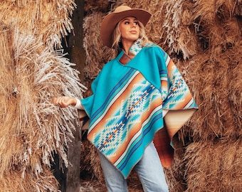 Könnte beinhalten: Eine Frau trägt einen türkisfarbenen und orangefarbenen Poncho mit einem braunen Cowboyhut und braunen Stiefeln. Sie steht vor einem Stapel Heuballen.