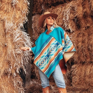 Puede incluir: Una mujer con un poncho estampado en turquesa y naranja, un sombrero vaquero marrón y botas marrones. Está de pie frente a una pila de balas de paja.