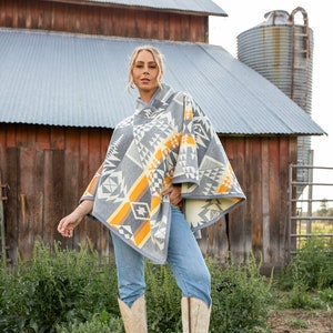 May include: A woman wearing a grey, yellow and white patterned poncho with a geometric design. She is wearing blue jeans and white cowboy boots. The woman is standing in a field of green grass.