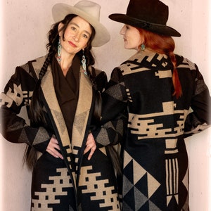 Può includere: Due donne indossano cappotti con motivi neri e marroni con colletti in pelliccia. La donna a sinistra indossa un cappello bianco e la donna a destra indossa un cappello nero.