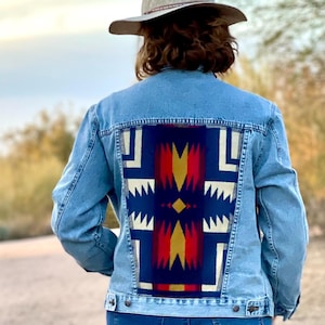 May include: Pale blue denim jacket with a vibrant geometric pattern on the back. The design incorporates red, yellow, and white elements against a dark blue backdrop. A light-coloured hat is worn.