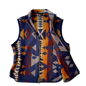 Può includere: Un gilet blu navy con un motivo geometrico in tonalità arancio, marrone e grigio. Il gilet ha una chiusura a zip e un colletto.