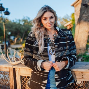 Può includere: Una donna indossa una giacca a righe bianche e nere con un motivo geometrico. Indossa jeans blu e una collana turchese.