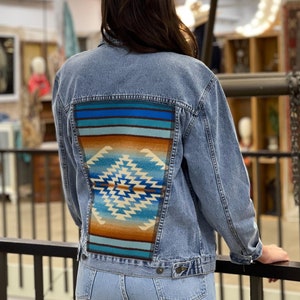 Könnte beinhalten: Eine hellblaue Jeansjacke mit einem farbenfrohen geometrischen Muster auf der Rückseite. Das Muster erinnert an einen indianischen Teppich oder eine Decke.