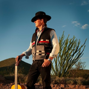 May include: A man wearing a black cowboy hat and a patterned vest stands in a desert landscape holding a wooden acoustic guitar.
