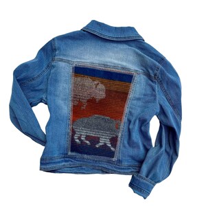 May include: A blue denim jacket with a colorful woven patch on the back. The patch features a stylized image of a buffalo and a landscape.