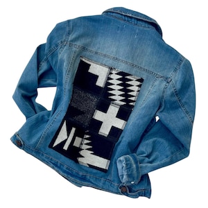 May include: Light blue denim jacket with a decorative patch on the back. The patch features geometric designs in black, white, and gray. The jacket has a classic collar, button closures, and pockets.