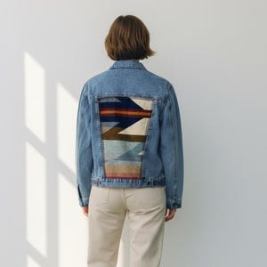 Puede incluir: Chaqueta vaquera azul claro con un diseño geométrico colorido en la espalda. La chaqueta se combina con pantalones beige y zapatillas blancas. El estilo general es casual y moderno.